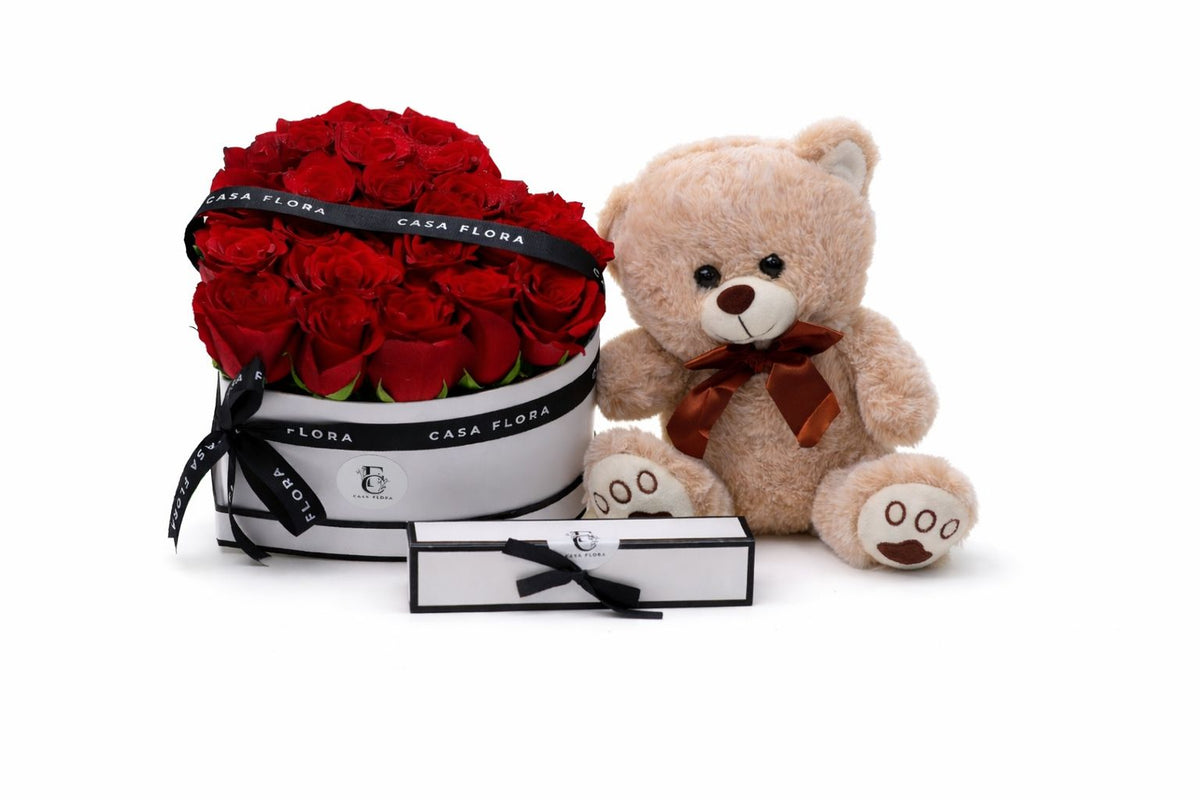 Heart-shaped box of red roses with a teddy bear and a card on a white background